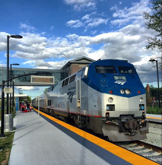 Front of Amtrak train arriving on a platform. #32 is on the front of the Silver and Dark Blue diesel locomotive. Glass walkway with information sign is above the train. 